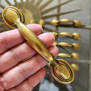 May include: Close-up of a brass-coloured cabinet pull with an oval backplate and a curved handle. The pull is held in a hand, with several other pulls visible in the background. The pulls have an antique or vintage aesthetic.