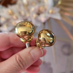 May include: Two shiny, gold-colored cabinet knobs with a round, ball-shaped design. The knobs are held in a person's hand, and the polished surface reflects the surrounding environment. The knobs are attached to a silver-colored screw.