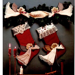 May include: Two red and white Christmas stockings with white lace trim and angel ornaments. The stockings are hanging from a garland of greenery with a paper scroll that says "Noel".