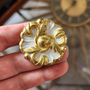 May include: An ornate, gold-coloured furniture fitting with white detailing. The piece has a floral design with intricate detailing. The hardware has a vintage aesthetic and is likely made of metal.