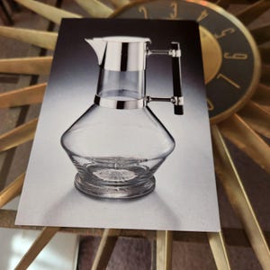 May include: A clear glass pitcher with a silver metal rim and handle. The pitcher has a conical shape with a wide base and a spout. The handle is black and silver. The pitcher is likely used for serving beverages.