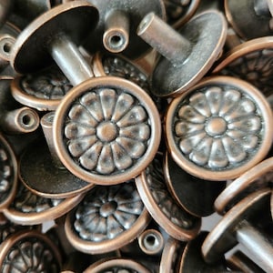 May include: Close-up of antique bronze cabinet knobs. Each knob features a round, floral design with a central button and a stem. The knobs are a warm, metallic brown colour and are piled together.