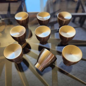 Peut inclure: Ensemble de neuf boutons de meuble de couleur bronze avec une finition en métal brossé. Les boutons ont un dessus arrondi et une base évasée, disposés sur une surface en verre. Pour meubles.