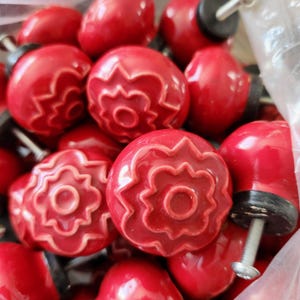 May include: A selection of red ceramic cabinet knobs with a floral design. Each knob has a glossy finish and a metal screw for installation. The knobs are round with a raised flower pattern and a central hole.