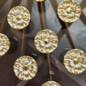 May include: A collection of circular buttons featuring a white floral relief pattern with metallic gold accents. Each button has a central gold dome surrounded by intricate, raised white petals and gold filigree details arranged in a symmetrical design.