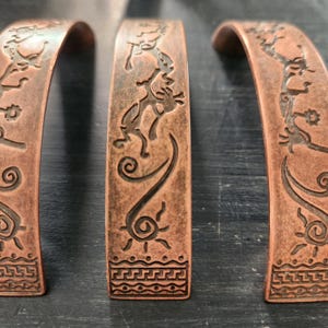 Peut inclure: Trois bracelets en métal couleur cuivre avec des motifs gravés. Les bracelets présentent des figures stylisées, des symboles solaires et des motifs géométriques. Le métal a un aspect chaud et vieilli, suggérant une esthétique artisanale ou vintage. Les motifs sont détaillés et complexes.