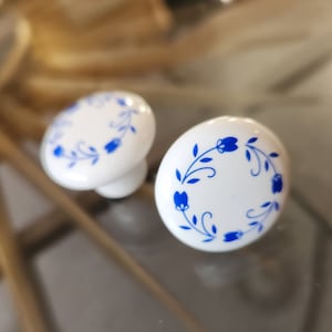 May include: Two white ceramic drawer knobs with blue floral designs. The knobs are round and have a raised edge.