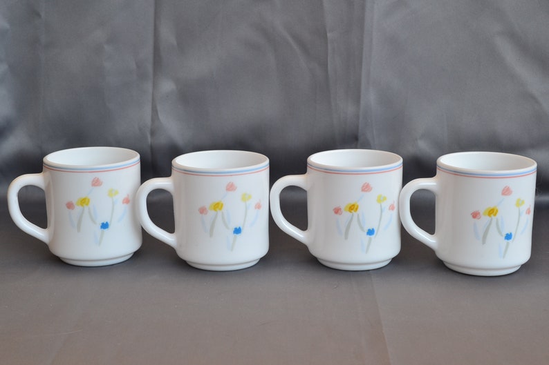 May include: Set of four white ceramic mugs with a floral design. The flowers are painted in shades of pink, yellow, blue, and green.