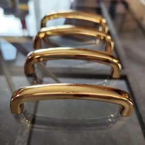 Peut inclure: Quatre poignées de porte de placard dorées avec une forme d'arche arrondie. Les poignées sont disposées en rangée, la plus grande à l'avant et la plus petite à l'arrière.