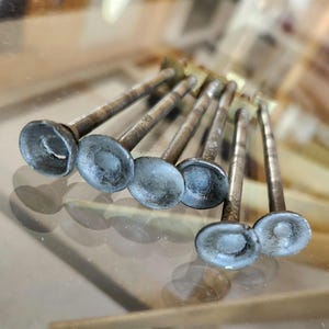 May include: A collection of vintage metal valve stems, each with a circular head and a long, slender shaft. The heads have a light blue hue, and the shafts show signs of age and wear. The items are arranged on a reflective surface.