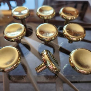May include: A collection of eight round cabinet knobs with a polished gold finish. Each knob features a smooth, domed top surface and a cylindrical base for mounting. The metallic surface reflects light, highlighting the circular shape and design.