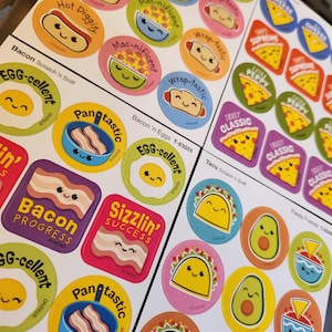 May include: A sheet of colorful food-themed stickers with smiling cartoon illustrations. Designs include a hot dog, egg, bacon, taco, avocado, and pizza. Text includes "Hot Diggity," "Egg-cellent," "Wrap-tastic," and "Truly Classic."