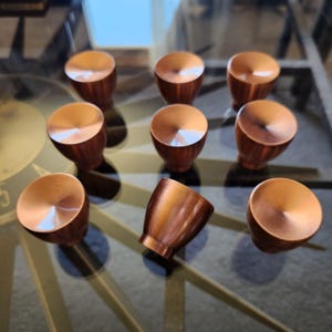 May include: A collection of nine copper-colored cabinet knobs. Each knob has a rounded top with a brushed finish and a tapered base. The knobs are arranged on a glass surface, reflecting the light and creating highlights.