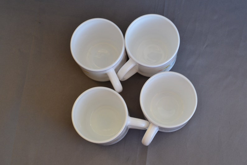 May include: Four white ceramic coffee mugs with a floral design. The mugs are empty and have a light brown background with a yellow and blue floral pattern.