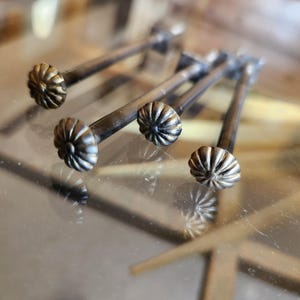 May include: Close-up of four antique bronze cabinet pulls. Each pull features a decorative, fluted knob and a slender, rectangular stem. The knobs have a detailed, floral-like design. The pulls are arranged diagonally across a reflective surface.