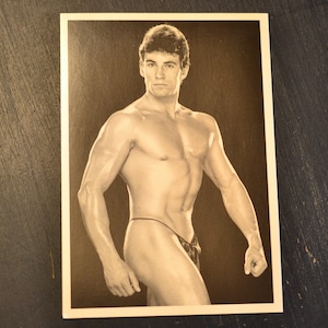 May include: Black and white photograph of a shirtless man with a muscular physique. He is standing with his left arm extended and his right arm bent at the elbow. He is wearing a thin black string around his waist.