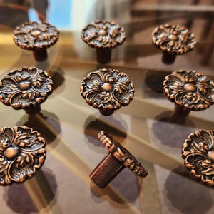 May include: Nine bronze-colored cabinet knobs with an ornate floral design. The knobs are round with a detailed pattern and a short stem. The knobs are arranged on a reflective surface.