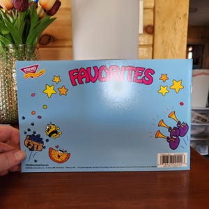 May include: A light blue book cover with the word "FAVORITES" in pink, yellow stars, and cartoon illustrations of bees, fruit, and purple characters playing trumpets. The book is held by a person.