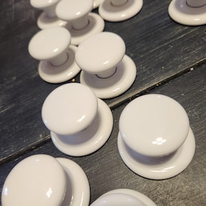 May include: A collection of white ceramic cabinet knobs with a rounded top and a circular base. The knobs are arranged on a dark wooden surface, showcasing their glossy finish and simple design. These knobs are suitable for furniture restoration or new builds.