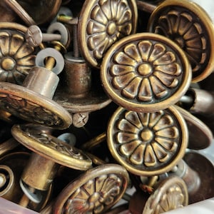 Peut inclure: Gros plan sur une collection de boutons de meubles anciens en laiton. Chaque bouton rond présente un motif floral détaillé et est fixé à une vis. Le laiton a un aspect vieilli et terni, suggérant une esthétique vintage.