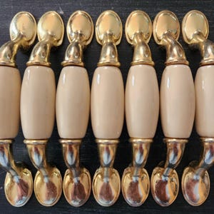 May include: A collection of eight cabinet pulls. Each pull features a cream-colored, oval-shaped handle with gold-toned metal accents. The pulls are arranged in a row, showcasing their decorative design and potential for furniture upgrades.