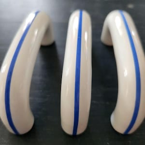 Peut inclure: Trois poignées de meubles en céramique blanche avec une finition brillante. Chaque poignée présente une seule bande bleue verticale au centre. Les poignées sont incurvées et ont une forme lisse et arrondie, adaptées aux tiroirs ou aux portes.
