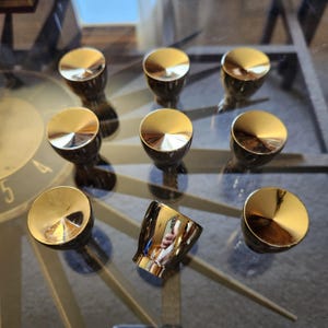 May include: A collection of eight gold-colored cabinet knobs with a reflective, polished finish. The knobs are arranged on a glass surface, reflecting the surrounding environment. One knob is tilted on its side.