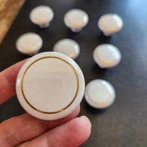May include: A collection of white ceramic cabinet knobs with a gold trim. One knob is held in a hand, while the others are scattered on a dark surface. The knobs have a classic, round design, suitable for furniture upgrades.