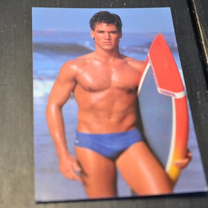 May include: A man with a muscular physique stands on a beach holding a red and yellow surfboard. He is wearing blue swim trunks.