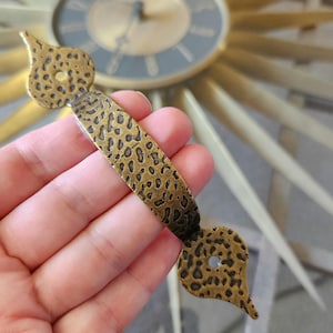 May include: A brass drawer pull with a hammered finish. The pull has a teardrop shape on each end and a smooth, slightly curved center section.