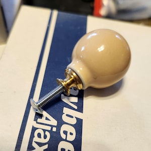 May include: A beige ceramic cabinet knob with a gold-coloured metal base and a silver screw. The knob is round and smooth, with a glossy finish. The screw is attached to the base of the knob.