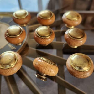 May include: Nine wooden cabinet knobs with brass-colored tops and bases are arranged on a glass surface. The knobs have a rounded shape with a natural wood grain finish. The brass accents add a touch of elegance to the hardware.