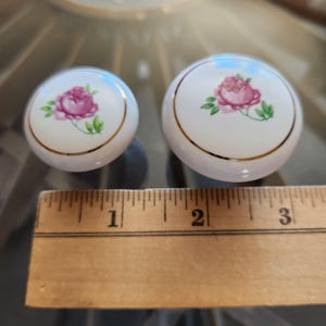 May include: Two round white ceramic cabinet knobs featuring a pink rose floral design with green leaves and a thin gold rim. A wooden ruler is positioned in front of the knobs, showing the smaller knob is approximately 1.25 inches in diameter.