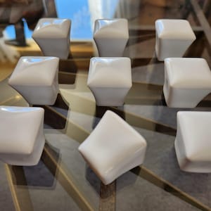 May include: Nine white ceramic cabinet knobs with a square top and a faceted design. The knobs are arranged on a glass surface with a geometric pattern. The knobs reflect light, creating highlights and shadows.