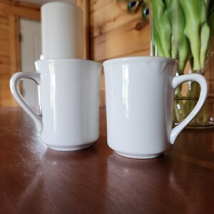 May include: Two plain white ceramic mugs with smooth, glossy finishes and curved handles. Each mug features a cylindrical body with a slightly flared rim and a sturdy base, presenting a clean, uniform appearance for everyday beverage consumption.