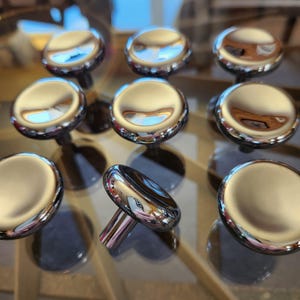 May include: A collection of eight circular cabinet knobs with a polished chrome finish. Each knob features a smooth, rounded top surface that reflects light, and a cylindrical base for mounting. The knobs are arranged in a grid-like pattern.
