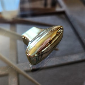 May include: A gold-colored, metallic object with a rounded, elongated shape and a rectangular top. The object reflects light, creating highlights and shadows. The item appears to be a decorative element or hardware component.