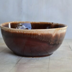 May include: A brown ceramic mixing bowl with a glossy finish. The bowl has a wide, rounded shape with a lighter brown rim. The exterior has a dark brown color that fades to a lighter shade near the rim.