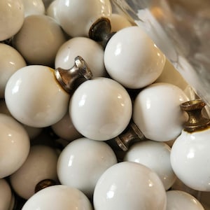May include: A collection of white, round cabinet knobs with a glossy finish. The knobs have a brass-colored base. The image shows a close-up view of the knobs, suggesting they are for furniture or decorative use.
