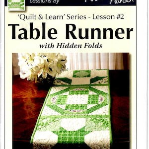 May include: A green and white table runner with a geometric pattern. The runner is made of fabric and has a quilted design. The text 'Quilt & Learn' Series - Lesson #2 Table Runner with Hidden Folds' is printed on the page.