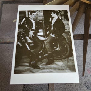 May include: Black and white photo of two men in leather jackets sitting on a motorcycle. The men are wearing jeans and have their hair slicked back. The motorcycle is a classic style with a large front wheel.
