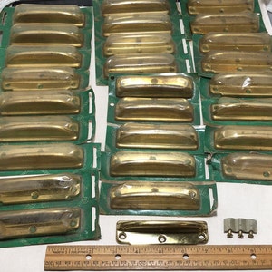 May include: A set of 18 brass cabinet pulls, each individually packaged in clear plastic. The pulls are rectangular with a slight curve and are packaged on green cards with the word "Amerock" printed on them. The pulls are approximately 7.6 cm long.