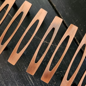May include: Close-up of several copper-colored, elongated metal handles with an oval cutout in the center. The handles are arranged diagonally on a dark, textured surface. The metal has a slightly aged appearance.