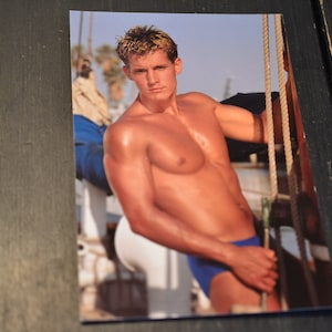 May include: A shirtless man with a muscular physique stands on a boat, holding onto a rope. He is wearing blue swim trunks and has a tanned complexion. The background is a blurry image of a boat and the ocean.