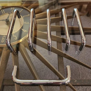 May include: A collection of shiny, chrome-finished cabinet pulls and handles. The hardware is arranged on a glass surface, reflecting the surrounding environment. The pulls have a curved, rectangular design, and are likely for furniture or cabinetry.