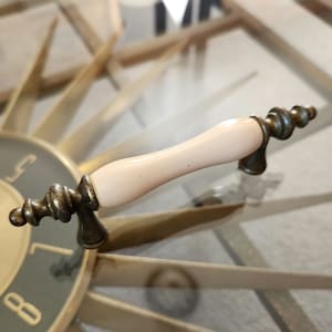 May include: A cream-colored ceramic cabinet knob with a brass base and a decorative scroll design.