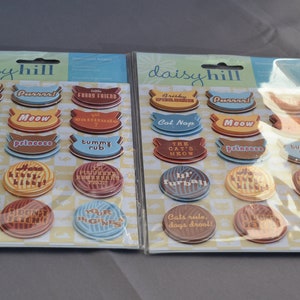 May include: Two packages of colorful, round stickers with cat-themed sayings. The stickers are on a white background with a checkered pattern. The packages are labeled "daisyhill".