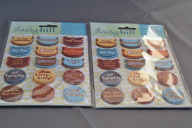 May include: Two packages of colorful, round stickers with cat-themed sayings. The stickers are on a white background with a checkered pattern. The packages are labeled "daisyhill".