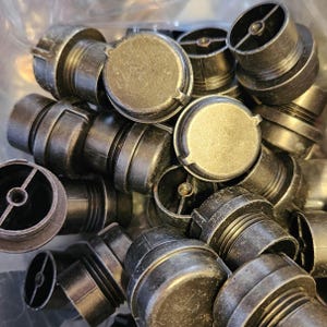 May include: A collection of dark gray plastic plugs with a metallic finish. The plugs have a circular top with a small tab and a threaded base. The image shows a close-up view of the plugs, with some partially visible.