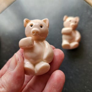 May include: Two small, light pink bear figurines. The bears are sitting, with their arms raised. The bears have small, dark eyes and a detailed snout. The figurines are made of a smooth material.